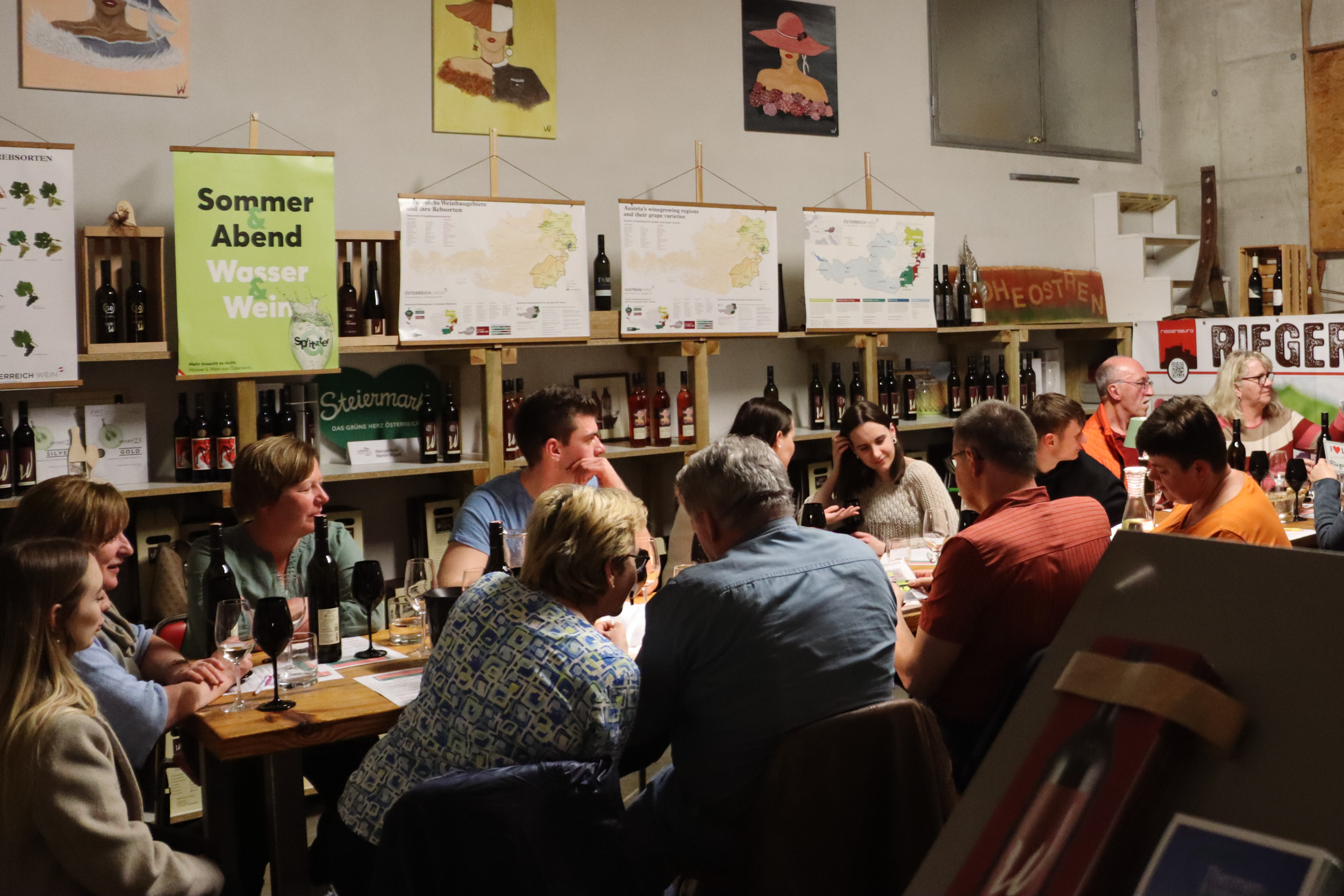 Eine Gruppe an Personen sitzt um eine lange Tafel. Am Tisch stehen gläserne und schwarze, undurchsichtige Weingläser. Im Hintergrund sind Regale voller Weinflaschen und Plakate mit Informationen rund um Wein zu sehen.