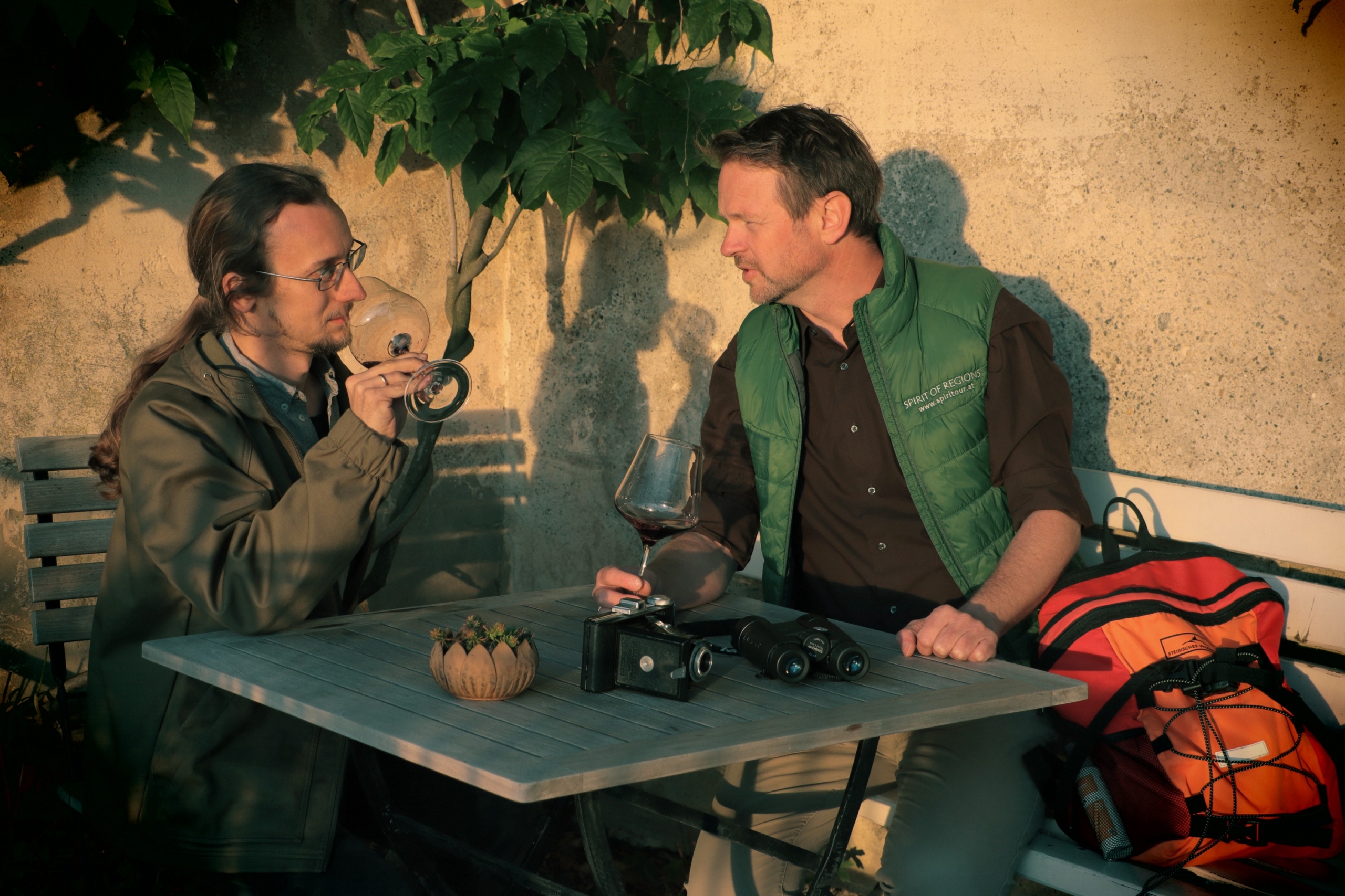 Zwei Männer sitzen im Abendlicht auf einer Bank und unterhalten sich bei einem Glas Weißwein