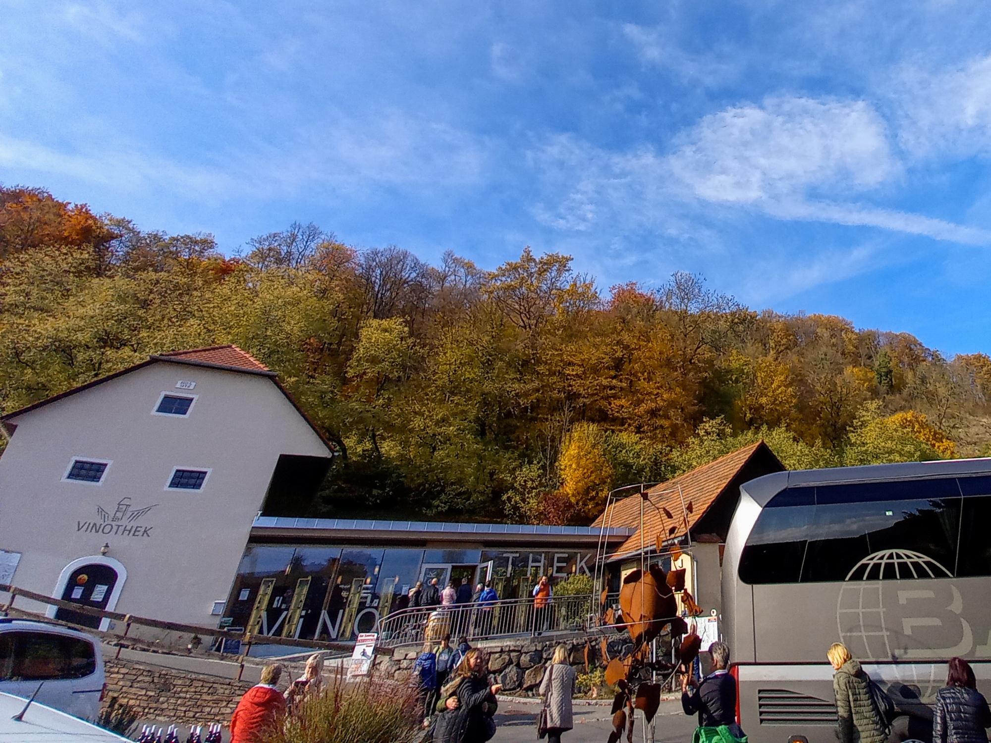 Vinothek Klöch Aus einem goldenen Bus steigen Personen aus und gehen über die Straße in ein Gebäude mit der Aufschrift Vinothek