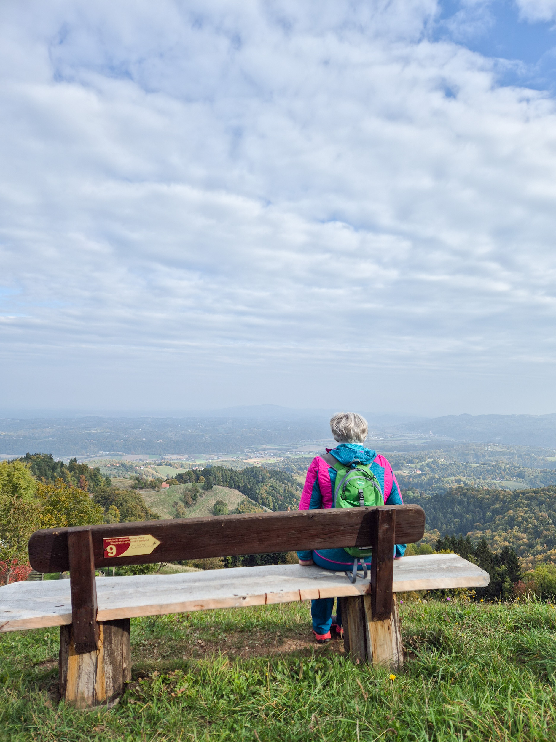 Wandern am Grenzpanoramaweg Eine Frau sitzt auf einer Holzbank und schaut von der Kamera weg auf das vor ihr liegende Panorama