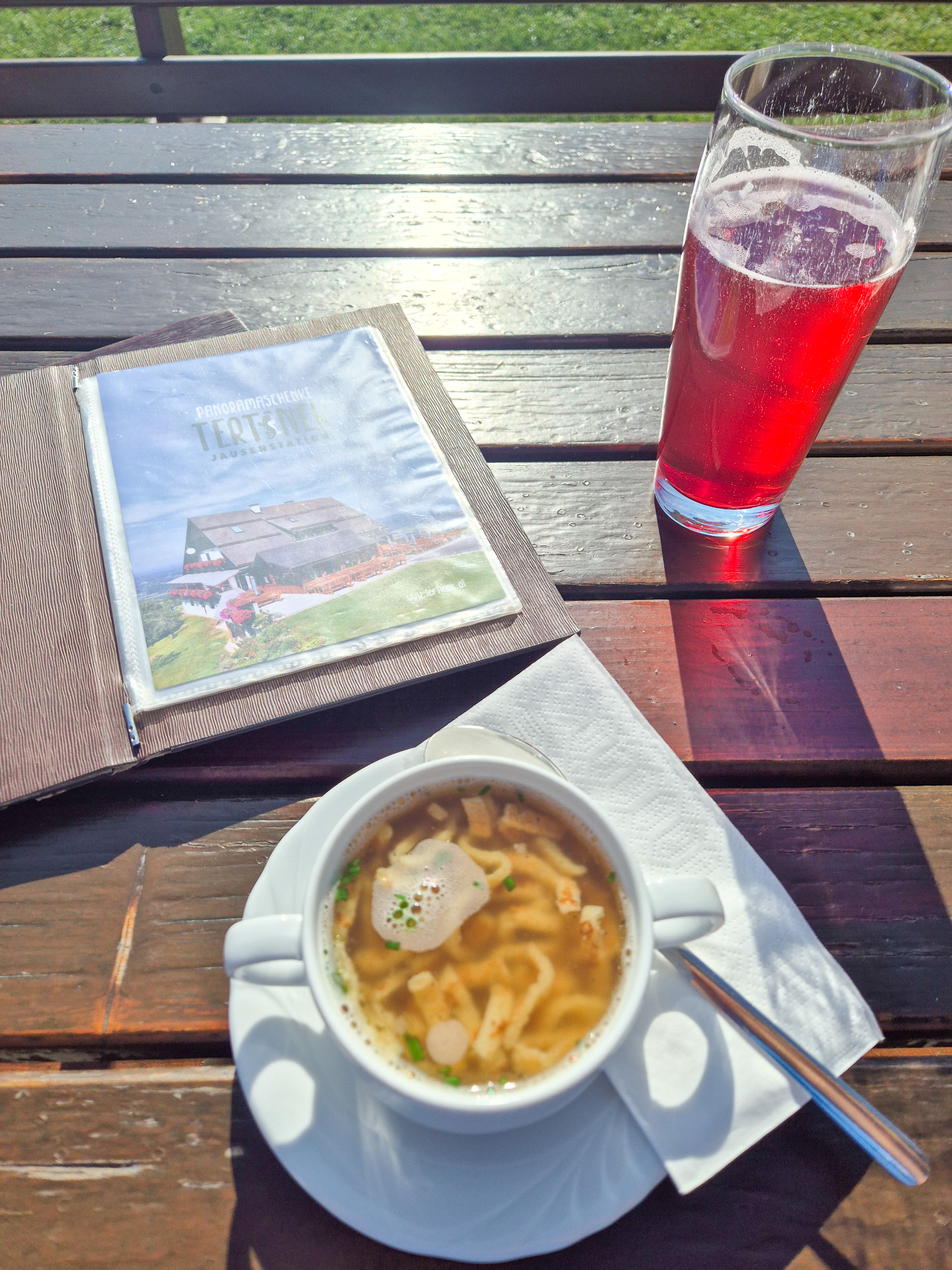 Wandern am Grenzpanoramaweg Auf einem Holztisch stehen eine Frittatensuppe, ein roter Saft und eine aufgeschlagene Speisekarte mit der Aufschrift Aussichtspunkt Tertinek