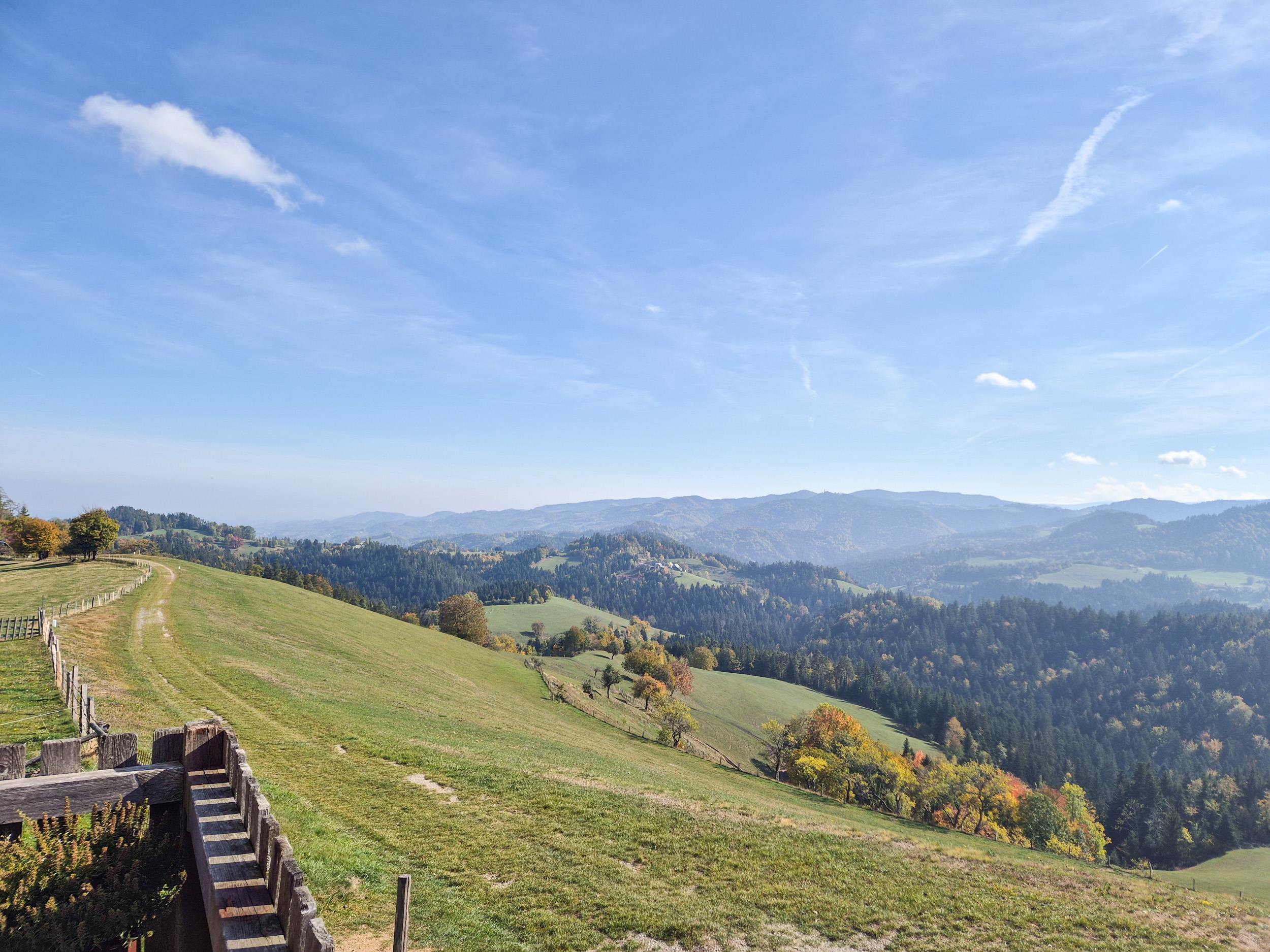 Wandern am Grenzpanoramaweg Von einer Holzterrasse blickt man auf den Wanderweg am Grenzkamm, dahinter sind slowenische Hügelketten sichtbar